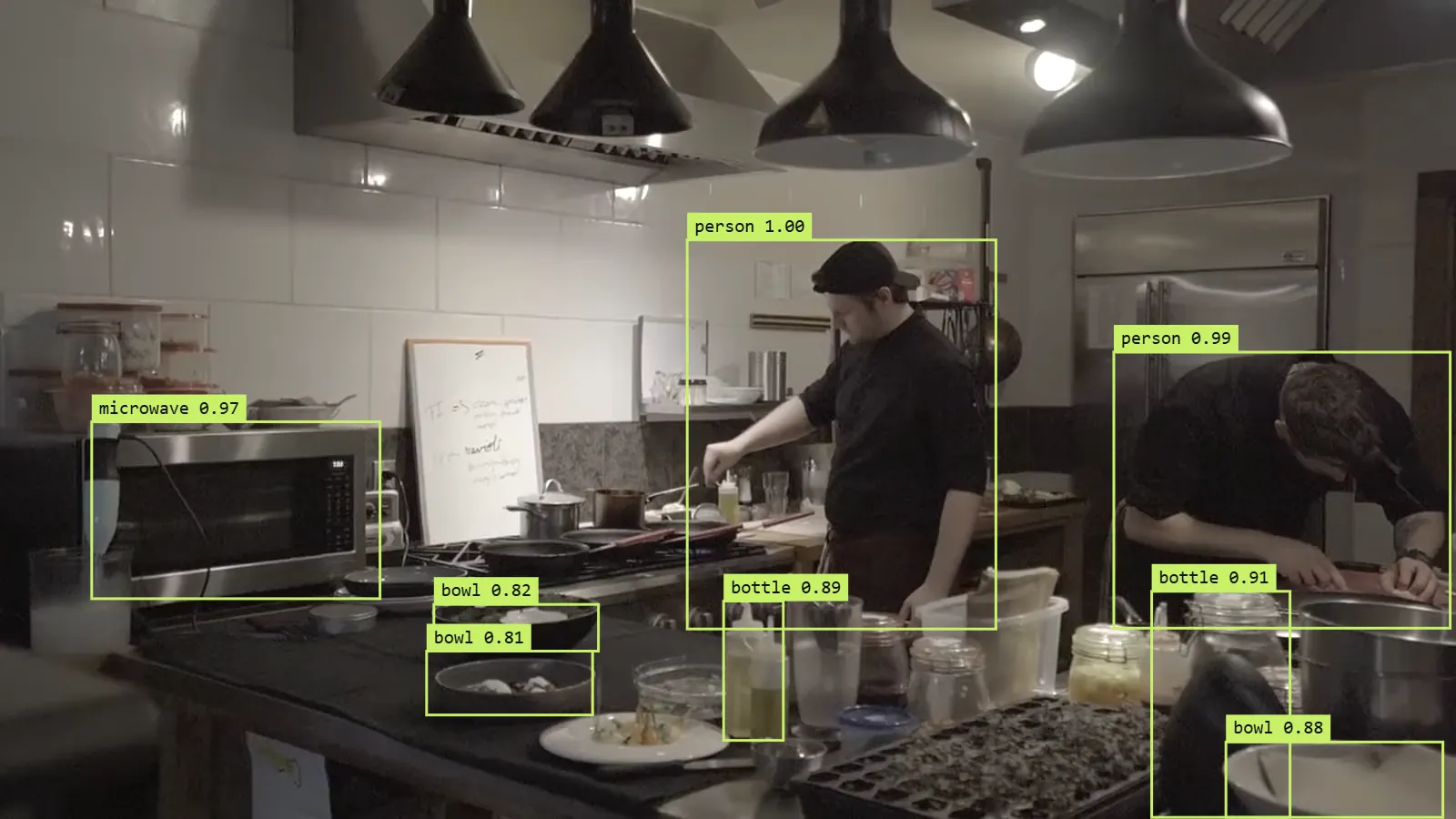 Frame of a kitchen with the detector boxes: two people, a microwave, bottles and bowls
